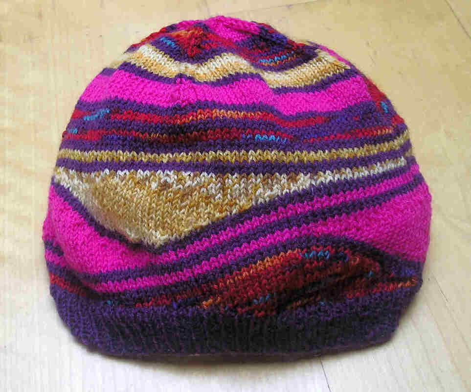 Gestrickte Damenmütze in Pink, Violett und Gold mit Rippenbund auf hellem Holzuntergrund