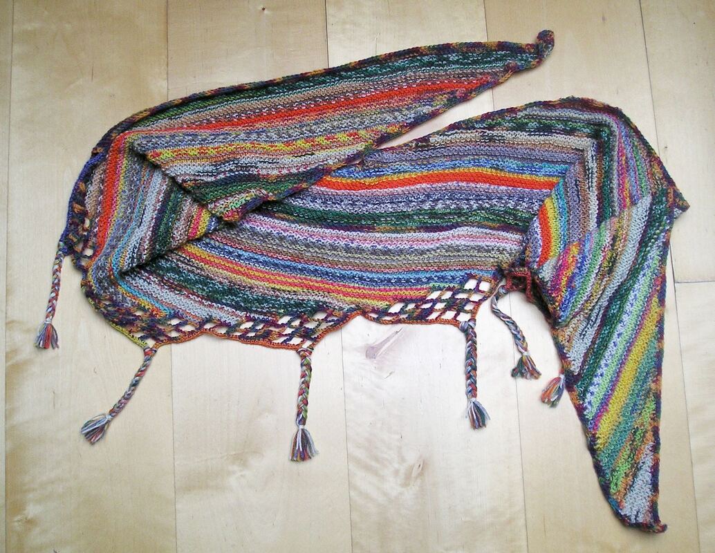 Mehrfarbiger gestrickter Dreiecksschal mit Streifen, Lochkante und Quasten auf hellem Holzboden.