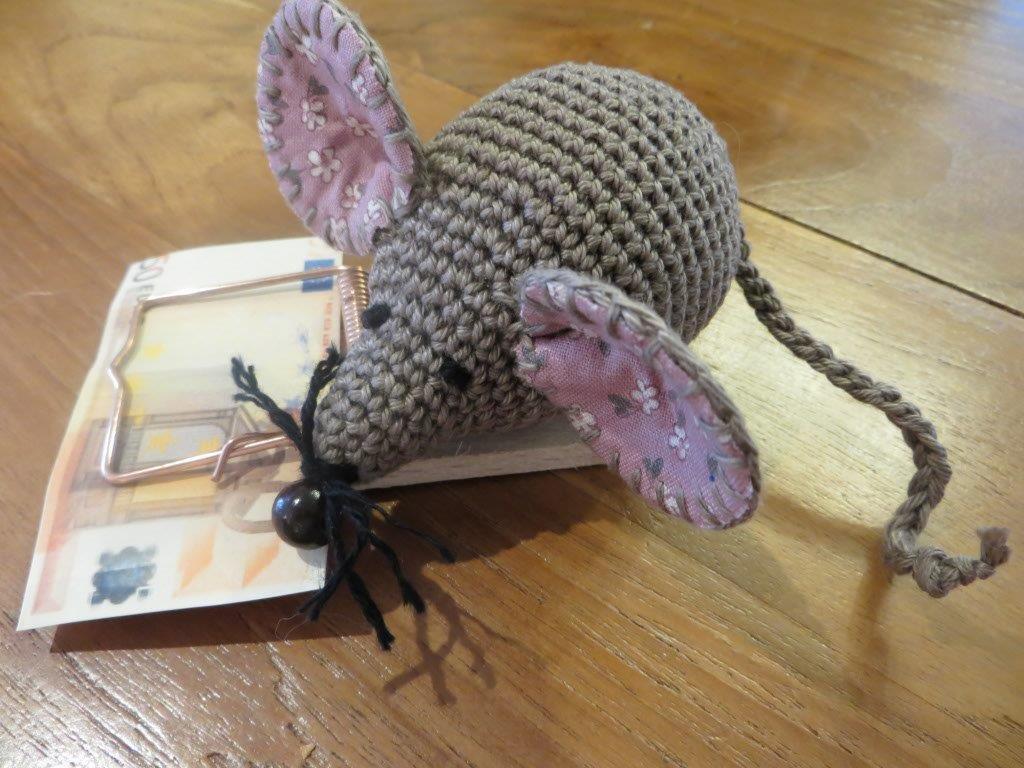 Gehäkelte grau-braune Amigurumi‑Maus mit rosa Stoffohren auf einer Holz‑Mausefalle, darunter ein Geldschein