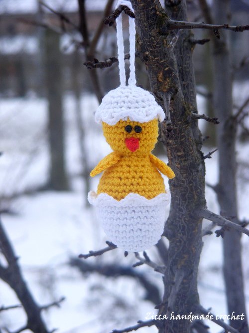 Gehäkelter gelber Küken-Anhänger in halb geöffneter weißer Eierschale mit Aufhänge-Schlaufe, hängend an einem Ast vor verschneiter Winterlandschaft