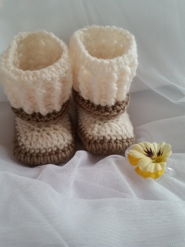 Zwei gehäkelte Babyschuhe in Creme und Beige mit umgeschlagenem Bündchen auf weißem Tüll, daneben eine gelbe Stoffblume