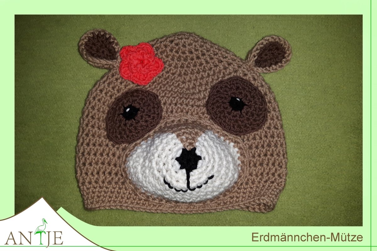 Gehäkelte Erdmännchen-Mütze mit runden Ohren, großen Augen, weißer Schnauze und roter Blüte auf grünem Stoff.