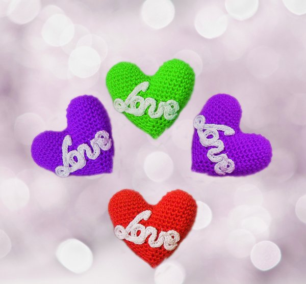 Vier gehäkelte Herzen in Rot, Grün und Lila mit weißer 'love'-Applikation vor rosa Bokeh-Hintergrund