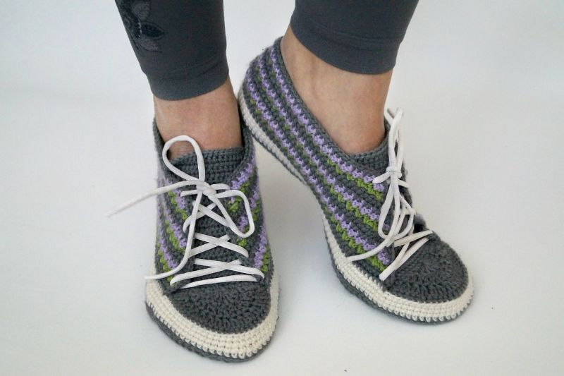 Füße in gehäkelten grauen Sneaker-Hausschuhen mit lila-grünen Streifen und weißen Schnürsenkeln