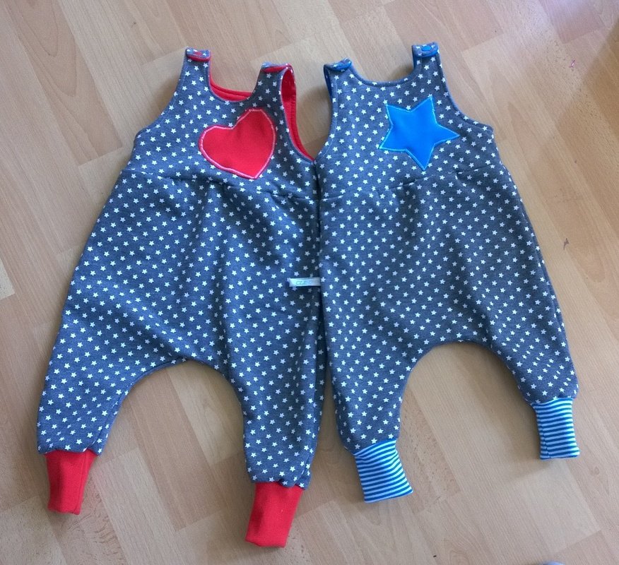 Zwei graue Babystrampler mit weißen Sternen, einer mit rotem Herz, einer mit blauem Stern, auf hellem Holzfußboden