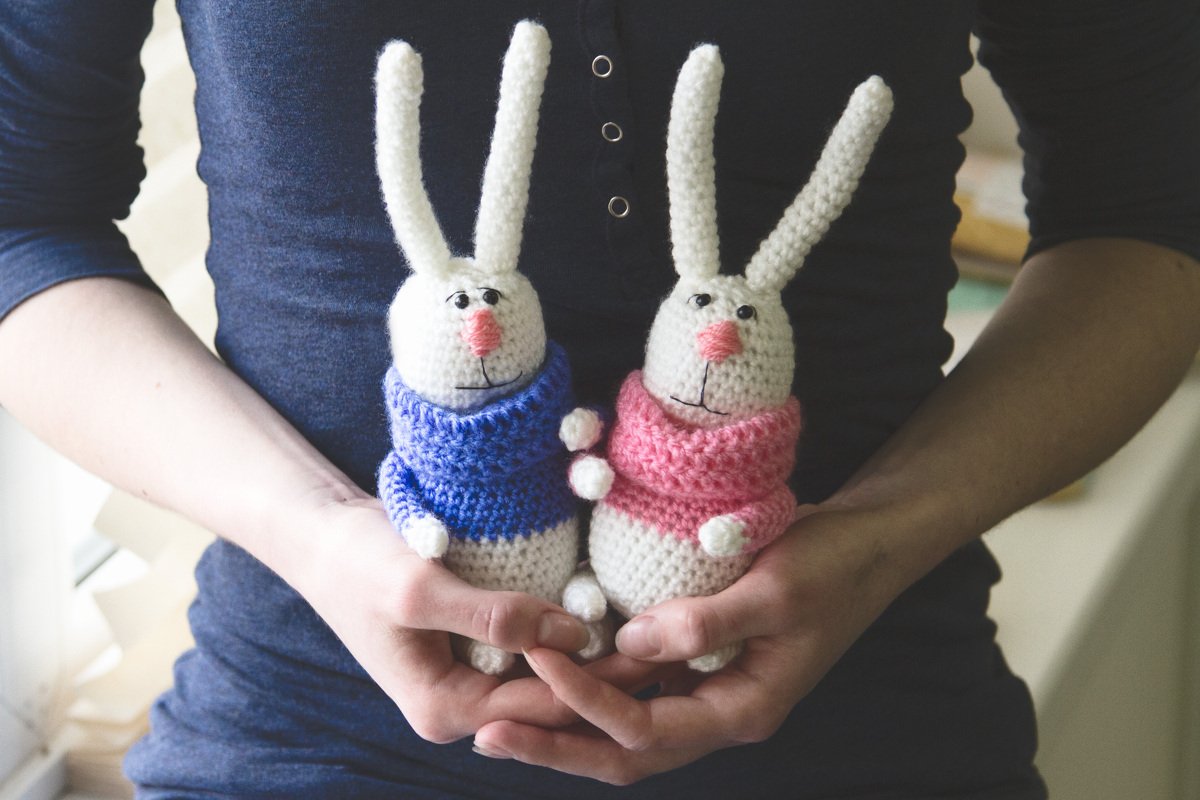 Zwei gehäkelte weiße Hasen-Amigurumi mit blauem und rosafarbenem Pullover in Händen
