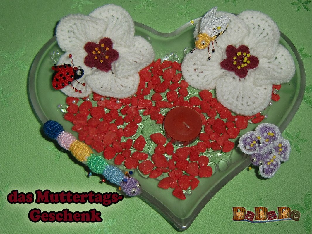 Zwei weiße Häkelblüten, gehäkelter Marienkäfer, Biene, Raupe und lila Blüten mit Teelicht auf Herzschale und roten Dekosteinen