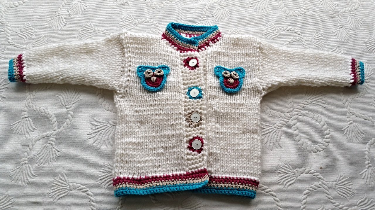 Cremefarbene, grob gestrickte Kinderjacke flach liegend mit mittiger Knopfleiste, türkis- und bordeauxfarbenen Kanten und zwei Eulen-Applikationen