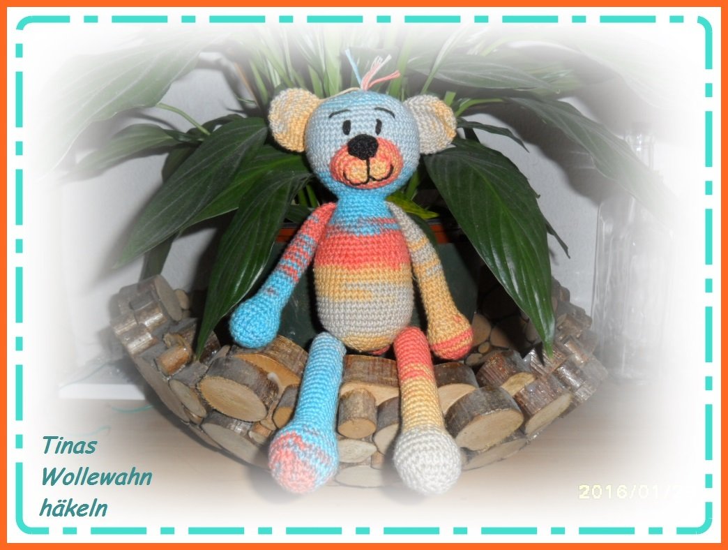 Mehrfarbiger gehäkelter Amigurumi-Teddy mit langen Beinen, sitzend in einer Holzschale vor einer Pflanze