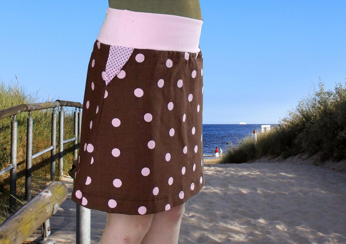 Brauner Kinderrock mit großen rosa Tupfen, hellrosa Rippbündchen und dreieckiger Tasche, vor Strandhintergrund