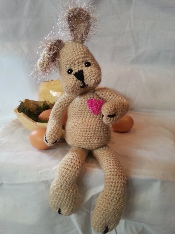 Beiger gehäkelter Osterhase mit rosa Herz neben Ostereiern und Schale mit Moos