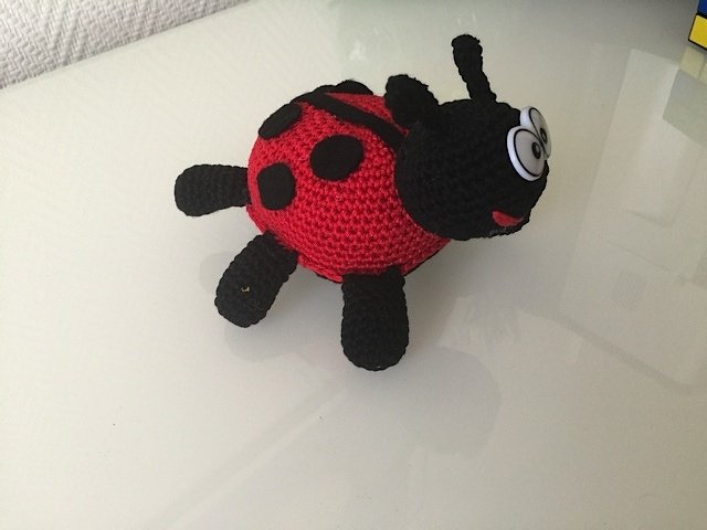 Gehäkelter Marienkäfer Amigurumi, rot mit schwarzen Punkten und großen Augen auf heller, glänzender Fläche.