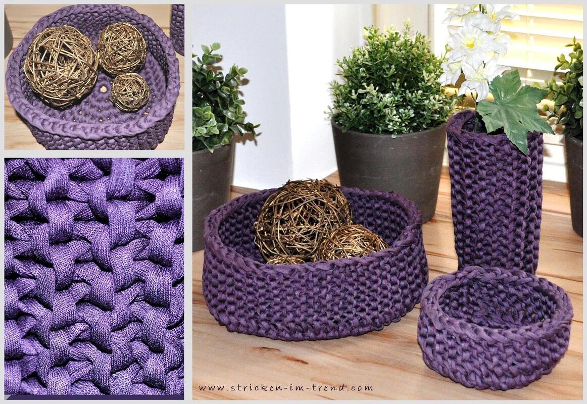 Mehrere violette, gehäkelte Körbe und ein hoher Behälter aus Textilgarn auf einem Holztisch neben Pflanzen.