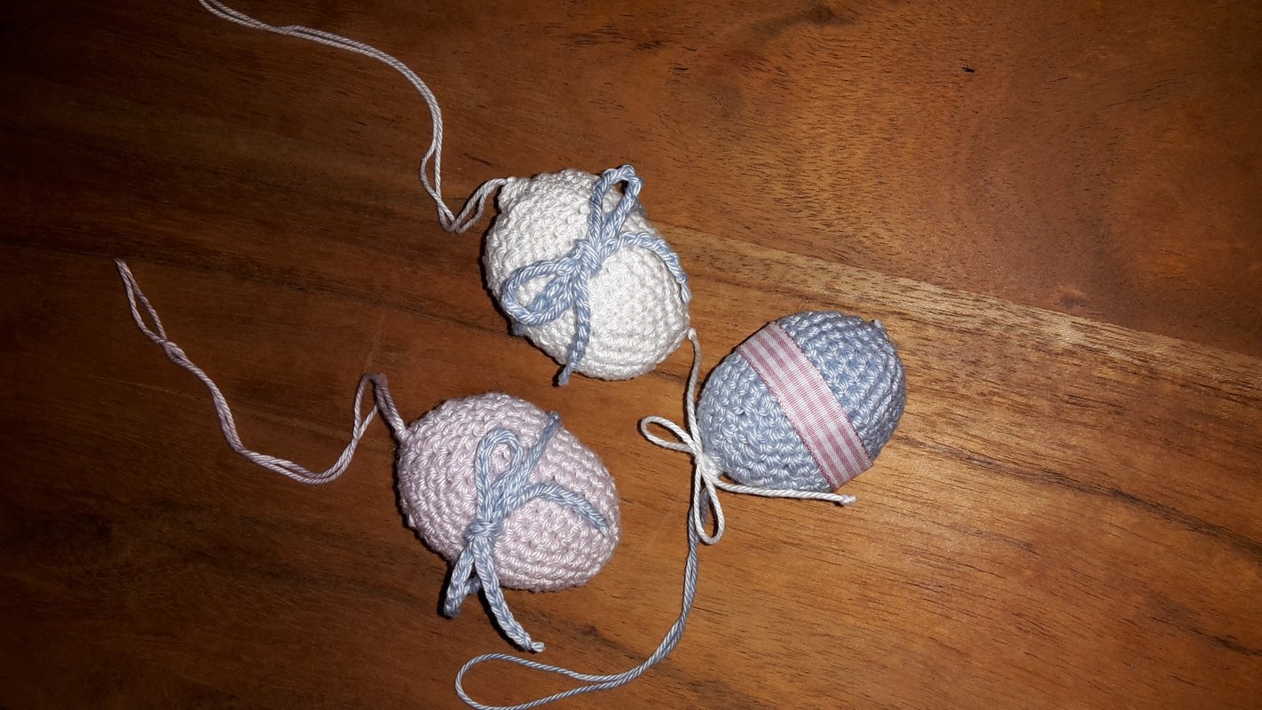 Drei gehäkelte Ostereier in Pastellrosa, Creme und Hellblau mit Schleifen auf Holzuntergrund