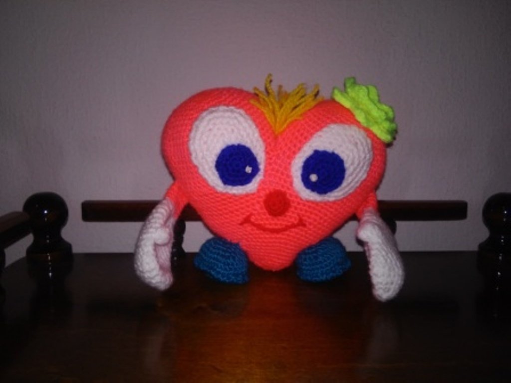 Korallenrotes Häkelherz-Amigurumi mit großen blauen Augen, Ärmchen und Füßchen auf einem Holztisch