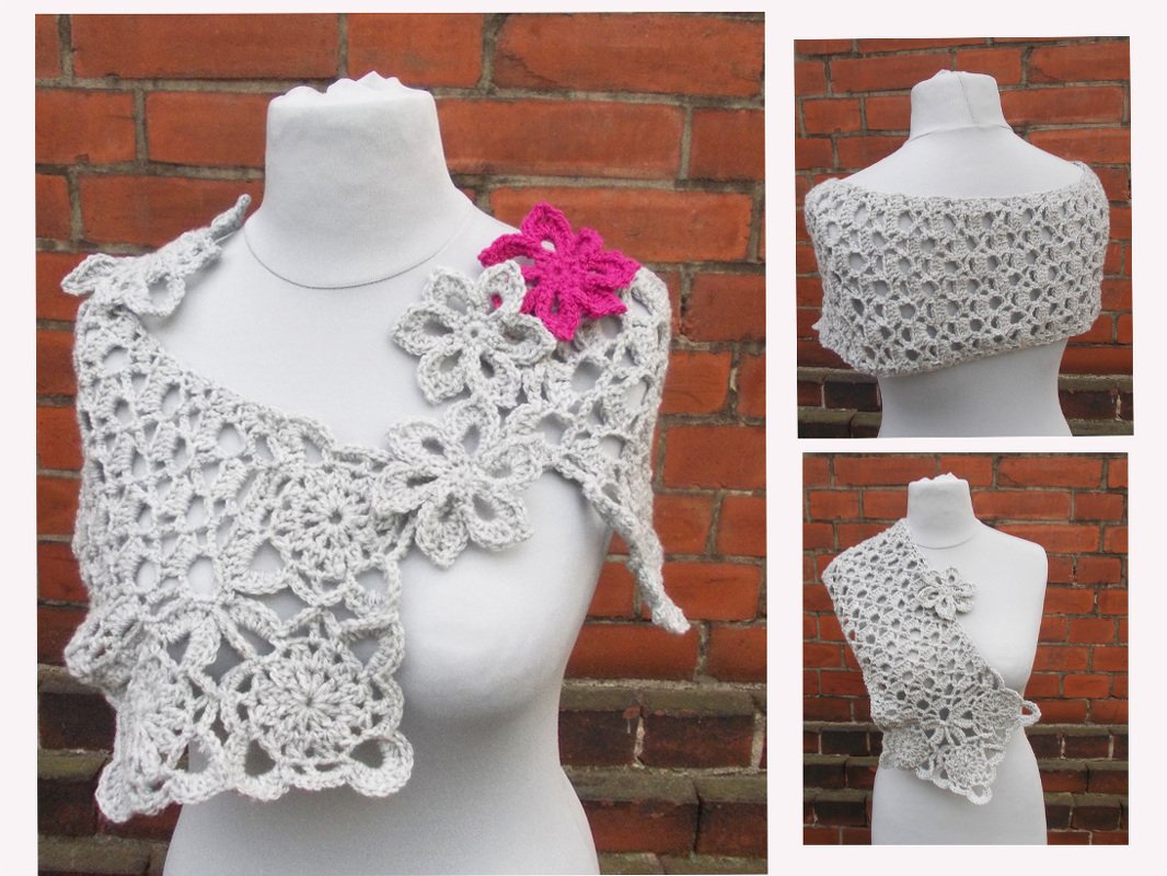 Gehäkelte Stola in hellem Grau mit Blüten- und Lochmuster, verziert mit einer pinken und mehreren grauen Blüten, auf weißer Schneiderpuppe vor Ziegelwand.