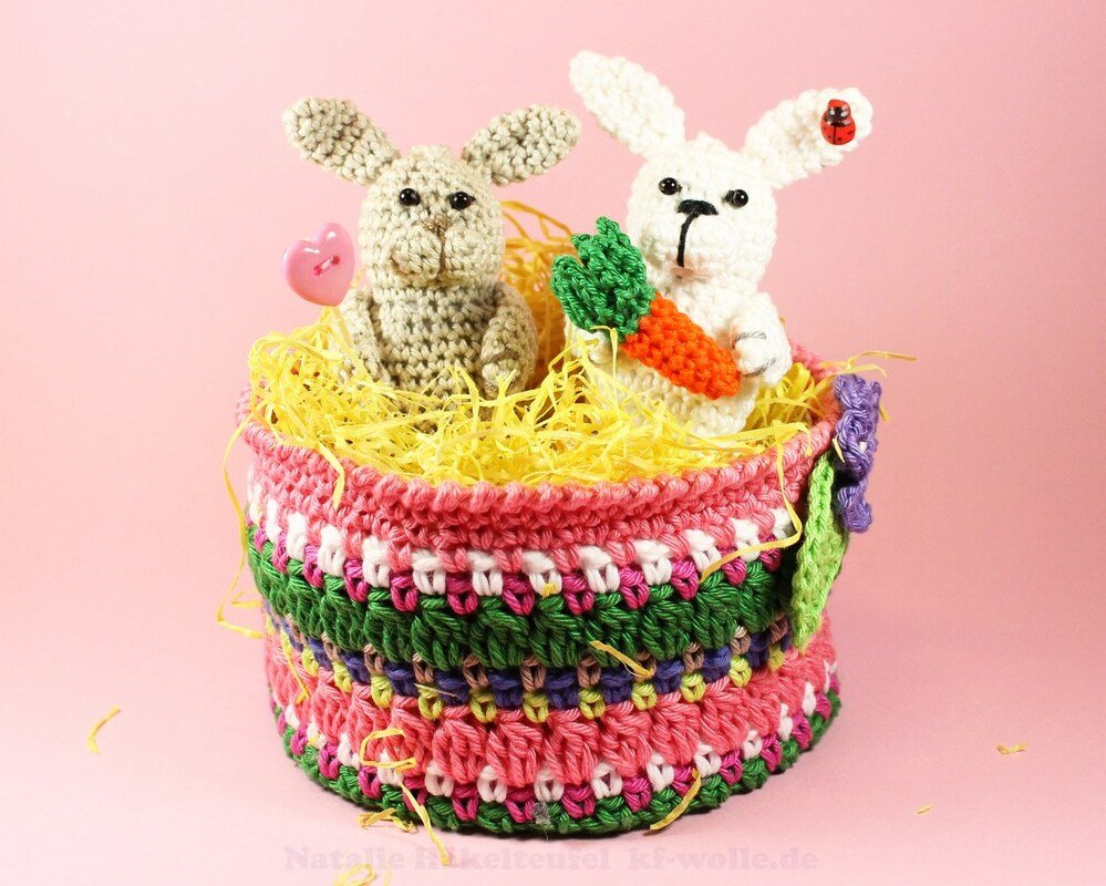 Zwei gehäkelte Amigurumi-Hasen in buntem Häkelkorb mit gelbem Ostergras vor rosa Hintergrund