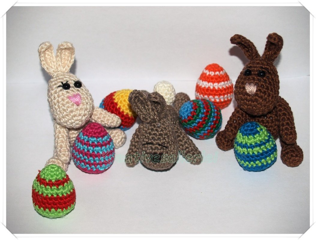 Drei gehäkelte Amigurumi-Hasen in Beige und Braun mit mehreren bunten, gestreiften Häkel-Ostereiern auf hellem Untergrund.