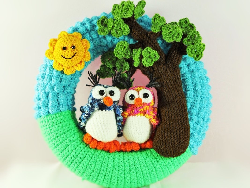 Crocheted wreath with two owls, a tree, and a smiling sun on blue sky and green ground.