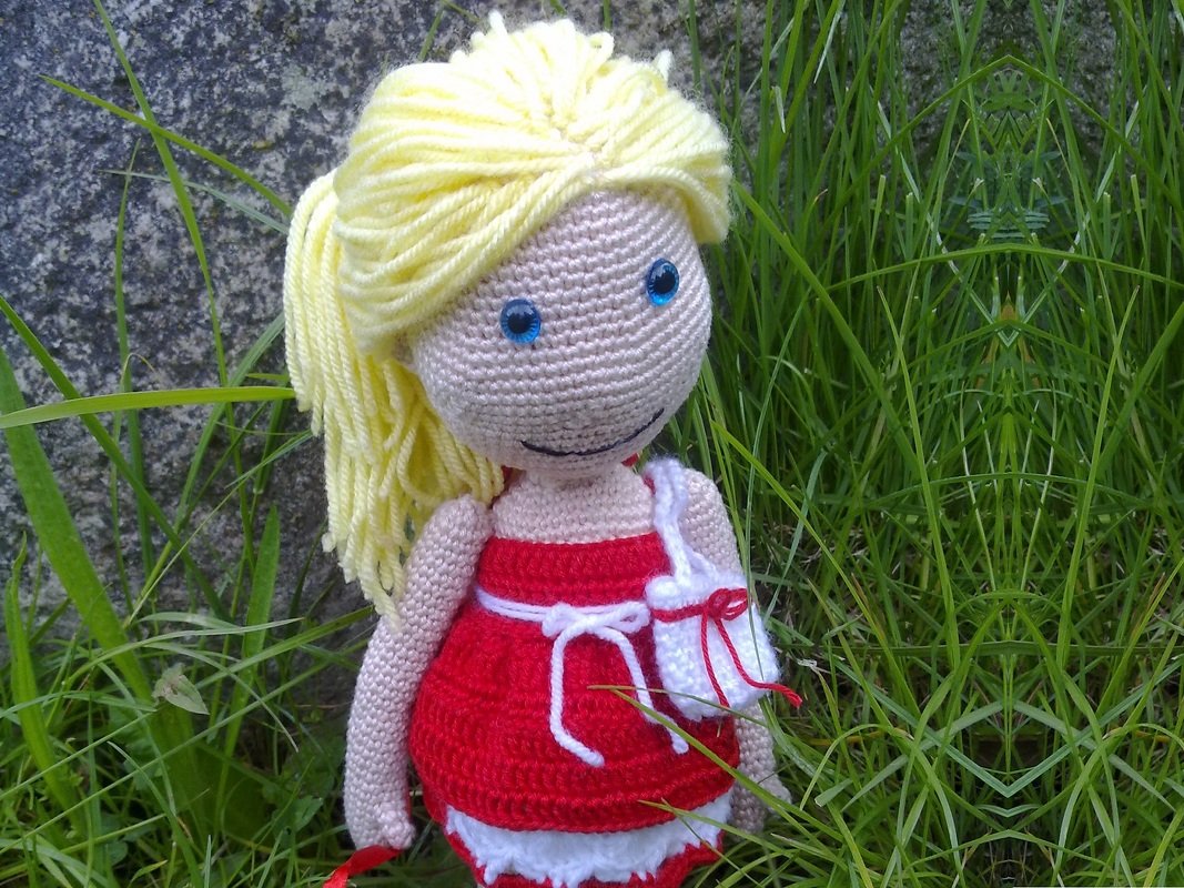 Detail einer gehäkelten Puppe mit gelben Garnhaaren, rotem Kleid und kleiner weißer Tasche im Gras
