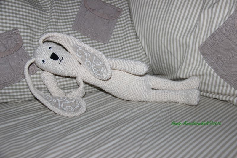 Cremefarbener gehäkelter Amigurumi-Hase liegt seitlich auf gestreifter Bettwäsche, lange Ohren mit weißer Stickerei sichtbar.