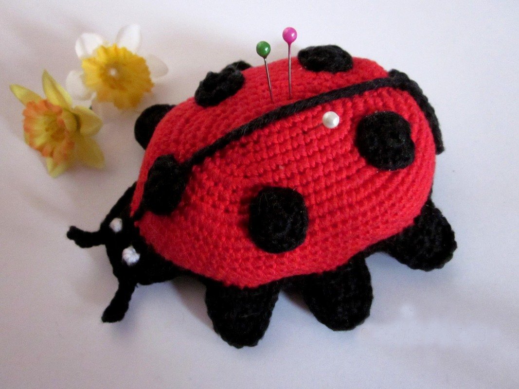 Gehäkeltes rotes Marienkäfer-Nadelkissen mit schwarzen Punkten und drei bunten Stecknadeln, daneben unscharfe Narzissen
