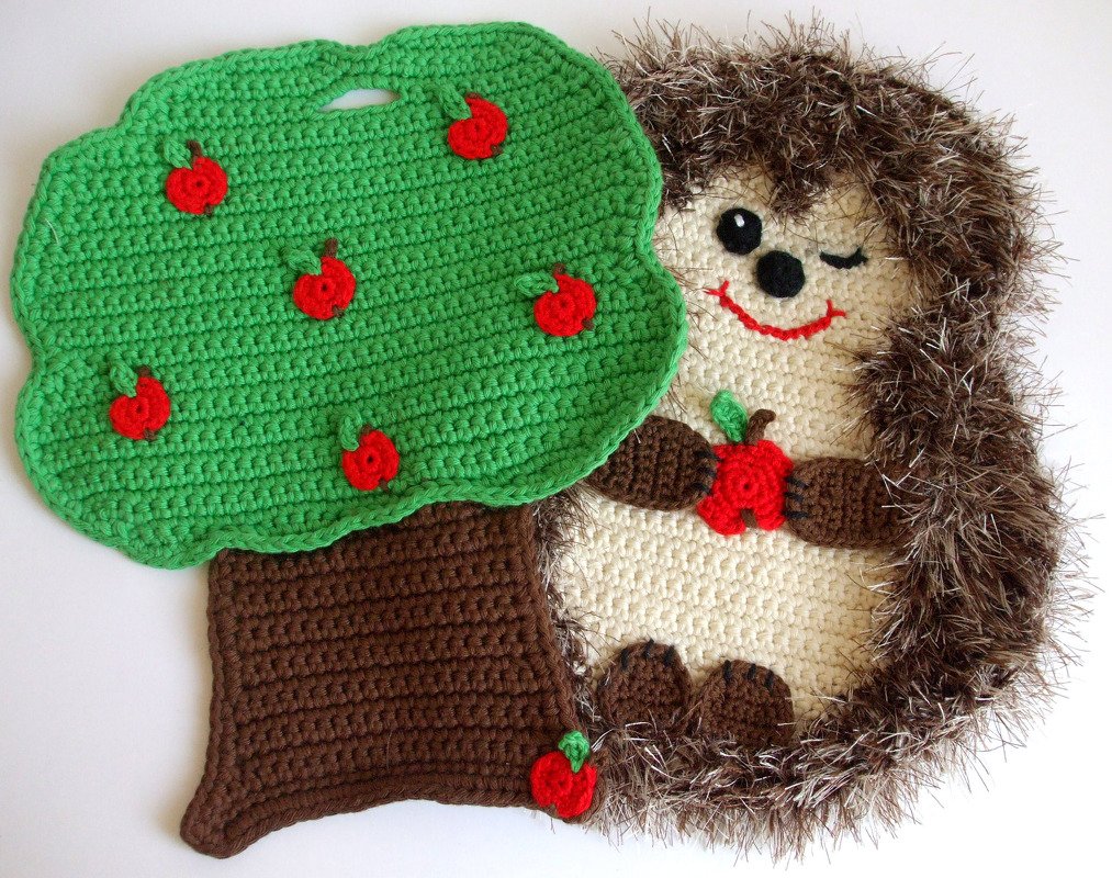 Gehäkelter Topflappen in Igel-Form neben gehäkeltem Apfelbaum-Topflappen mit roten Äpfeln und Griffschlitz