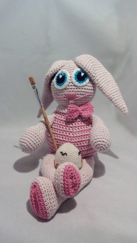 Gehäkelter rosa-weißer Amigurumi-Hase mit blauen Augen und rosa Fliege, der einen Pinsel hält; ein kleines gehäkeltes Osterei sitzt vor ihm.