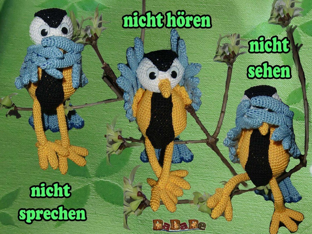 Drei gehäkelte Amigurumi‑Vögel in Blau, Gelb und Schwarz auf Zweigen in den Posen nicht hören / nicht sehen / nicht sprechen