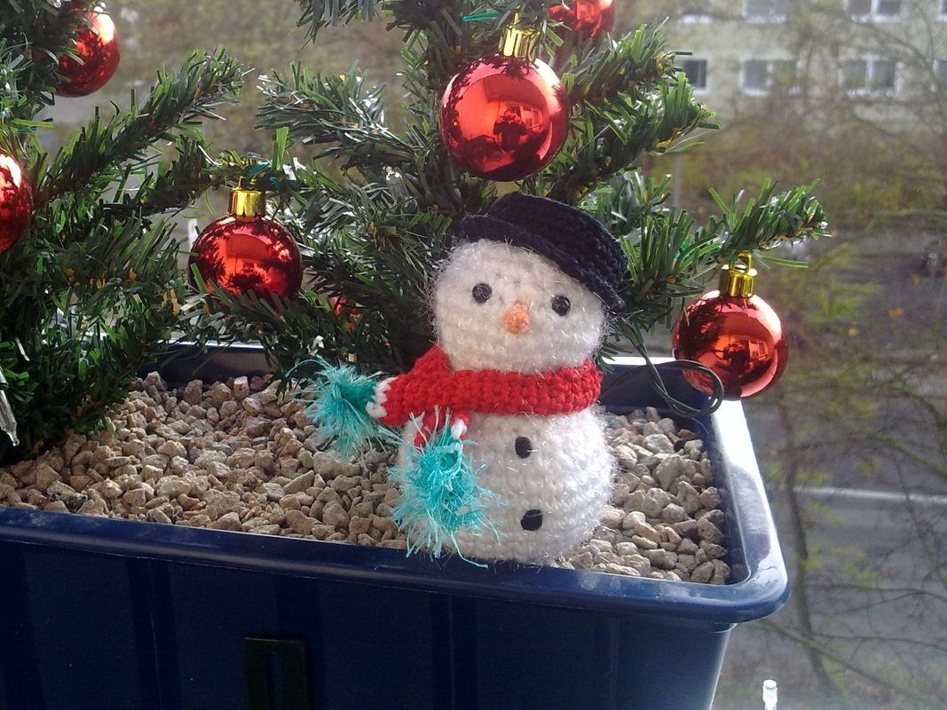 Gehäkelter Amigurumi-Schneemann mit Hut und Schal im Blumenkasten neben künstlicher Tanne und roten Weihnachtskugeln