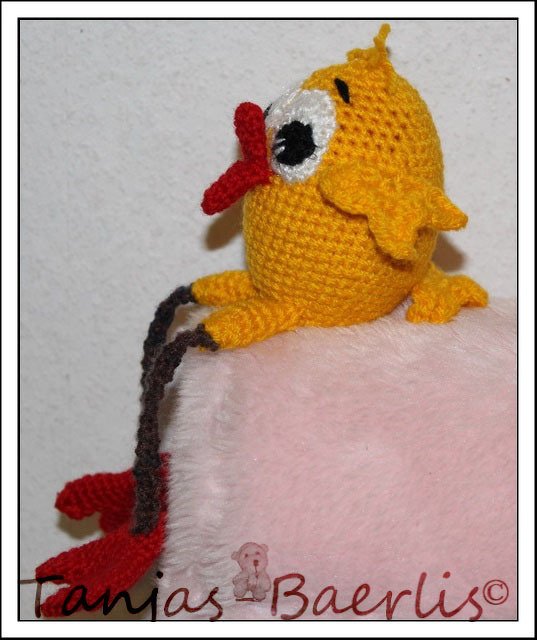 Gehäkeltes gelbes Küken mit rotem Schnabel und roten Füßen auf rosa Unterlage.