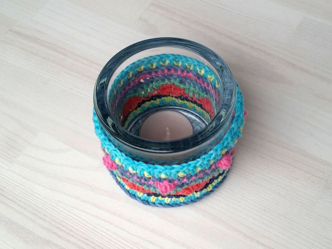 Glas-Teelichthalter mit gehäkeltem Überzug in Türkis, Pink und Orange auf hellem Holztisch, Teelicht im Inneren.