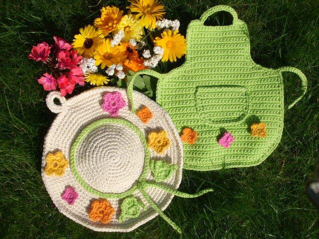 Zwei gehäkelte Topflappen auf Gras: runder beiger Untersetzer mit Blumen und grüner Topflappen mit Tasche