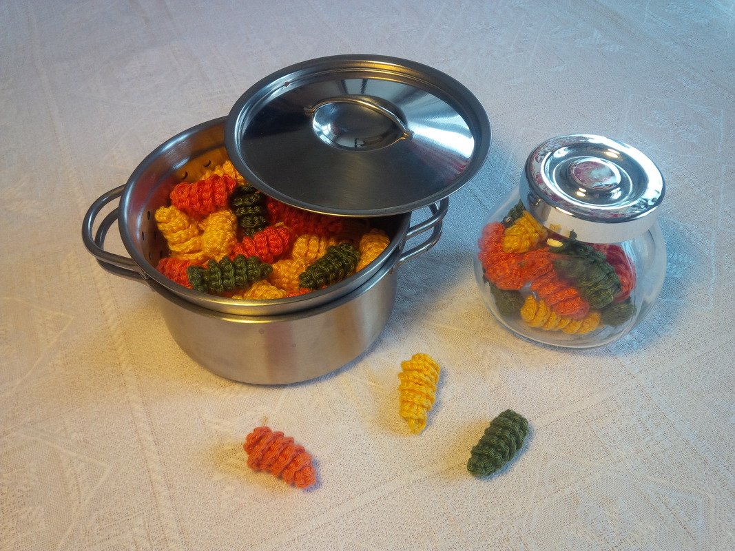 Bunte gehäkelte Nudeln in einem Topf mit Siebeinsatz, in einem Glas und lose auf einer Tischdecke