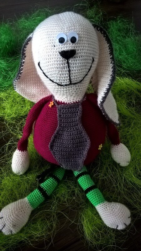 Gehäkelter Amigurumi-Hase mit weißem Kopf, langen Ohren, dunkelrotem Körper, grauer Krawatte, gestreiften Beinen und Wackelaugen auf grünem Dekogras.