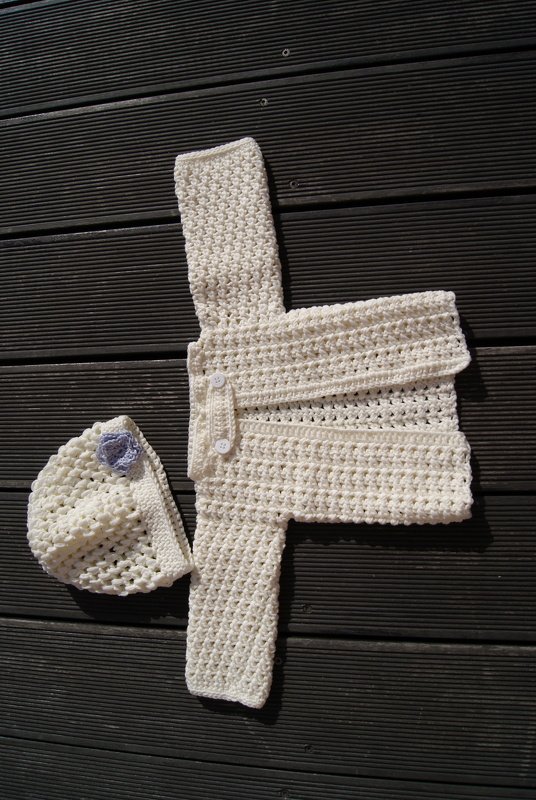Cremefarbenes gehäkeltes Babyjacken-Set mit Lasche und Knöpfen sowie Mütze auf Holzuntergrund