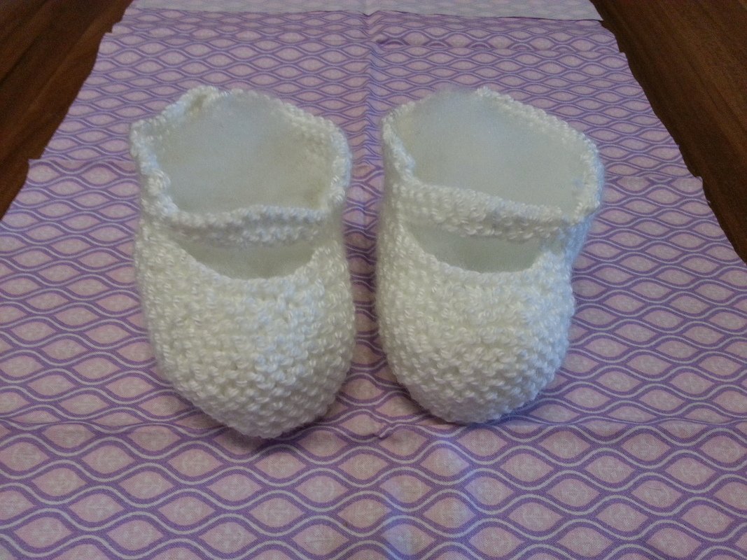 Paar weißer handgestrickter Babyschuhe mit Riemchen auf lila Stoff