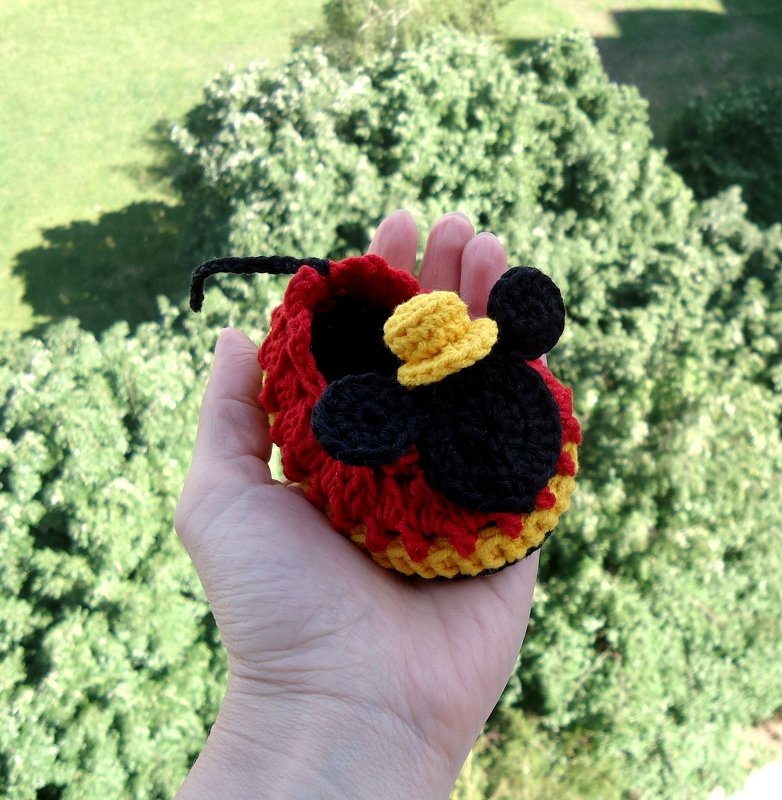 Adult hand holding a red and yellow crocheted baby bootie with black ear-shaped applique and small yellow hat