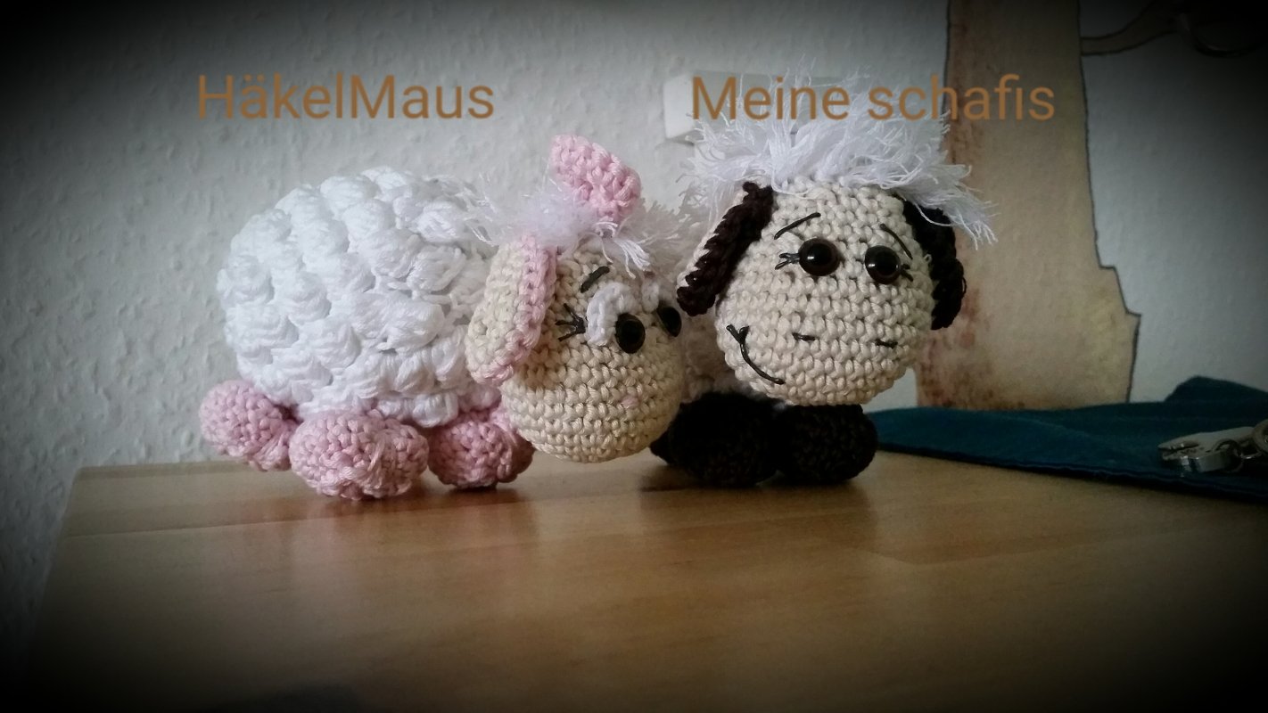 Zwei gehäkelte Amigurumi-Schafe, eines weiß-rosa, eines beige-braun, sitzend auf einem Holztisch.