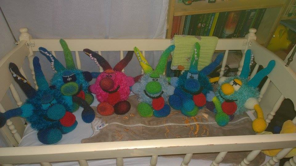 Fünf bunte, gehäkelte Amigurumi-Monster mit unterschiedlichen Gesichtern und Gliedmaßen liegen auf einer hellen Decke in einem weißen Kinderbett.
