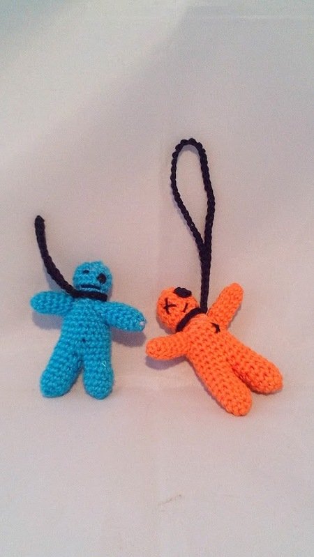 Zwei gehäkelte Amigurumi-Anhänger, einer türkis, einer orange, mit gestickten Gesichtern und schwarzen Aufhängeschlaufen auf hellem Hintergrund.