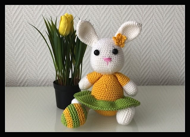 Weißes gehäkeltes Amigurumi-Häschen im gelb-grünen Kleid mit gestreiftem Osterei neben einer Tulpe im Topf