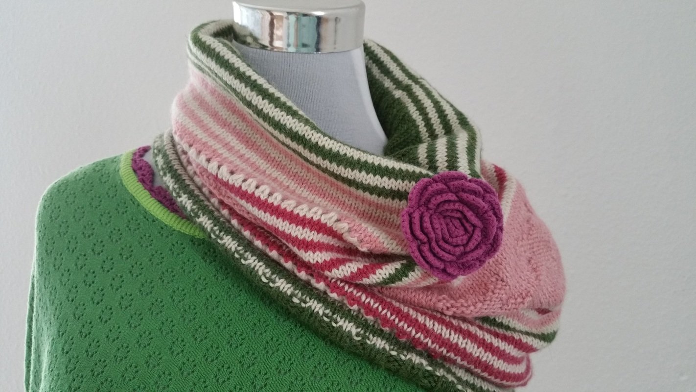 Gestrickter Loop-Schal in Grün, Rosa und Creme mit gestreiftem Muster und violetter Blumenbrosche auf Schaufensterpuppe
