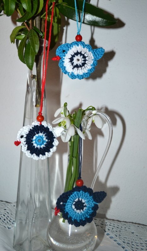 Drei gehäkelte Vogelapplikationen in Blau- und Weißtönen mit roten Perlen, dekorativ an Zweigen und in Vasen mit Schneeglöckchen.