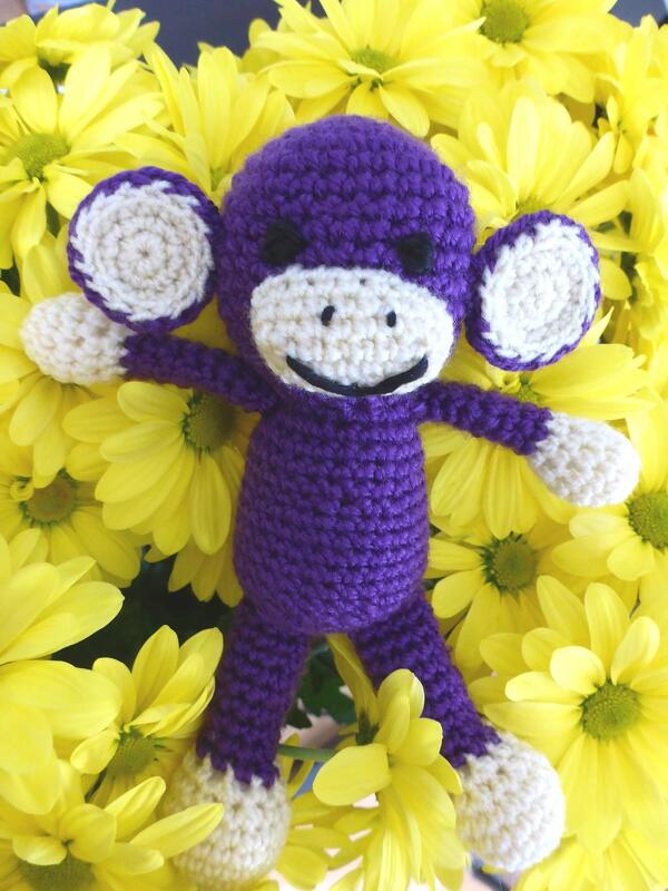 Gehäkelter lila Amigurumi-Affe mit cremefarbenem Gesicht und Gliedmaßen auf gelben Blumen