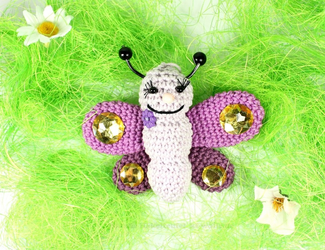 Gehäkelter Amigurumi-Schmetterling mit hellviolettem Körper und goldfarbenen Schmucksteinen auf grünem Dekogras.