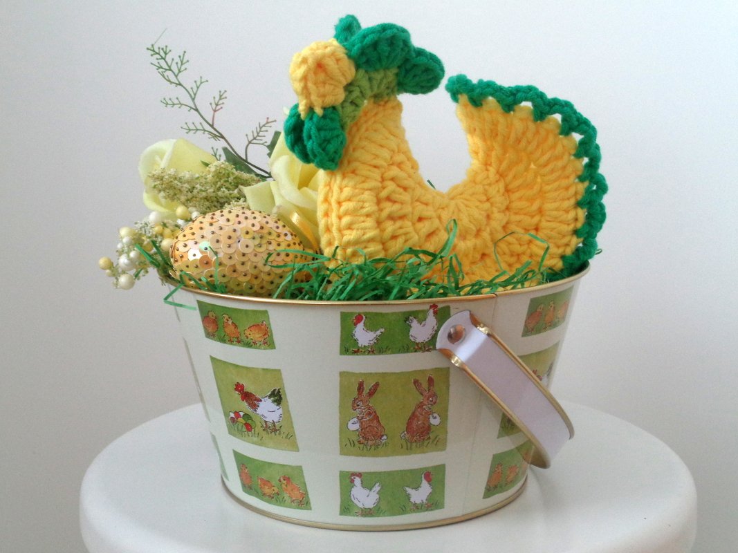 Gehäkeltes gelb-grünes Huhn in einem dekorativen Eimer mit Osterei und Blumen