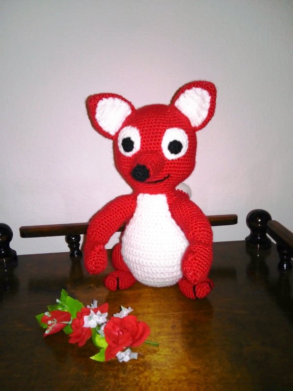 Rotes und weißes gehäkeltes Amigurumi-Tier mit großen Ohren auf Holzoberfläche, rote Blumen im Vordergrund