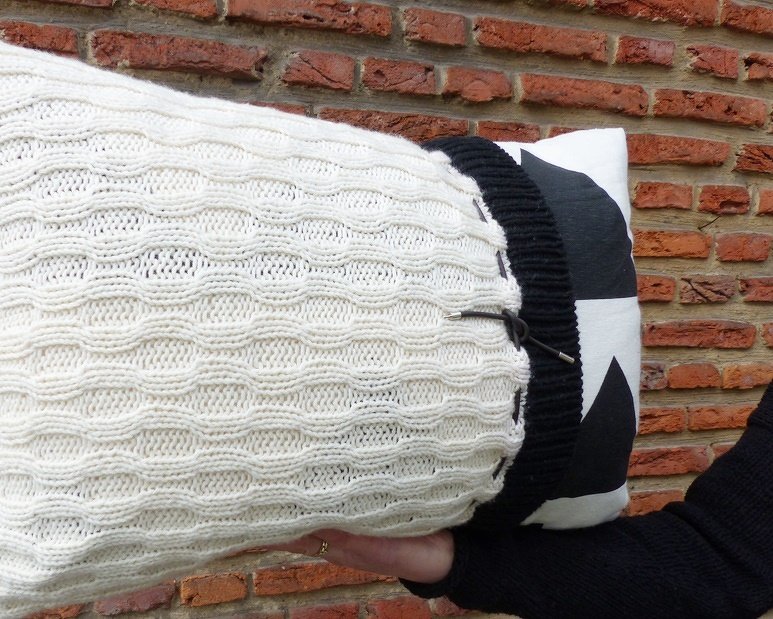 Weißer gestrickter Rollenkissenbezug mit Wellenstruktur und schwarzem Bündchen vor Ziegelwand