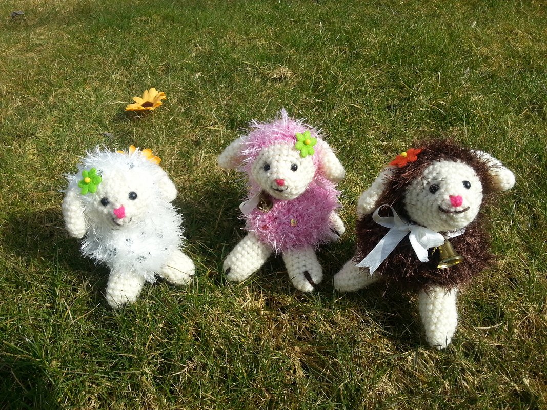Drei gehäkelte Amigurumi-Lämmer in Weiß, Rosa und Braun auf Gras mit Blüten, Schleife und Glocke